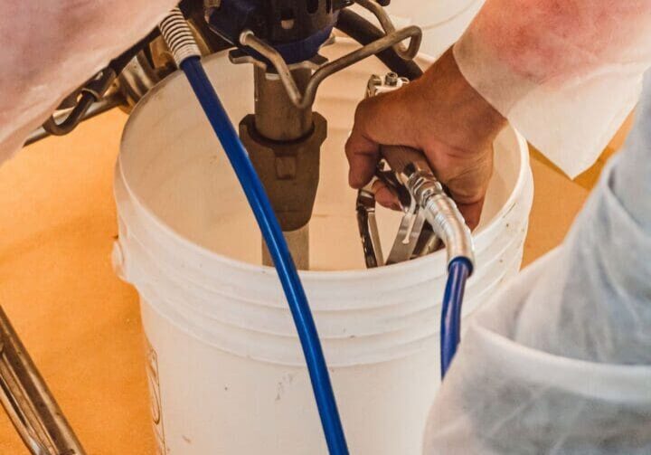 DSC03851 Person in protective clothing operates a spray paint machine over a white bucket, holding a blue hose and gun in hand.