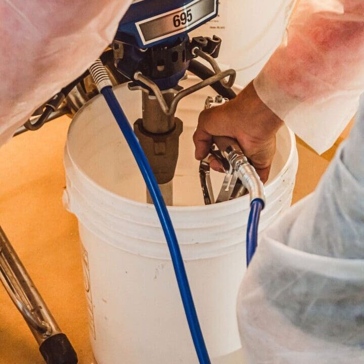 DSC03851 Person in protective clothing operates a spray paint machine over a white bucket, holding a blue hose and gun in hand.