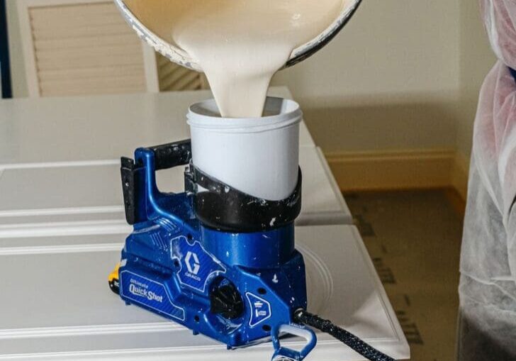 DSC04020 Paint being poured from a container into a blue paint sprayer, with a lid and hose nearby on a table.
